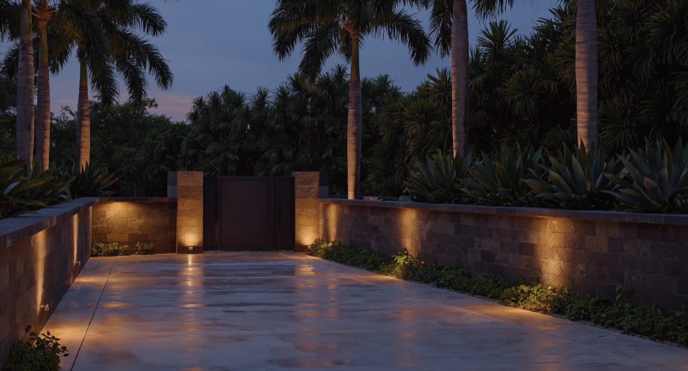 Artist's impression of an estate drive at dusk: a lit stone wall, a
                  private gate between stone piers, and palms behind.